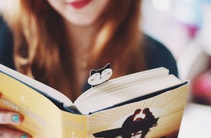 A girl is reading a book with a cat bookmark in it.