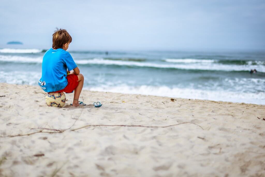 A boy is looking at the ocean while sitting on a soccer ball.