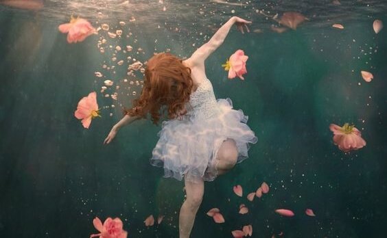 Woman in dress floating underwater surrounded by flowers.