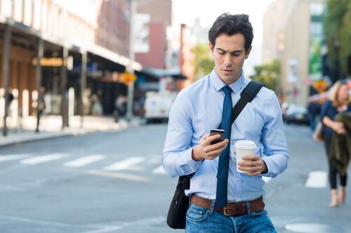 A man in a shirt and tie walking down the street with a coffee in one hand and his phone in another.