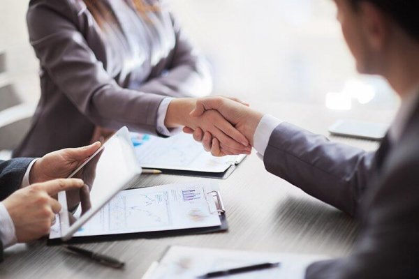 Two people shaking hands during an interview while another person is using a tablet.