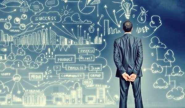 A person in a suit seen from the legs up standing in front of a board with various illustrations and flow charts drawn digitally on it.