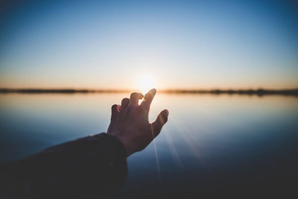 A person's hand reaching out towards a sunset over the water. 