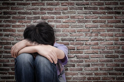 A boy sitting against a brick wall with his face in his arms.
