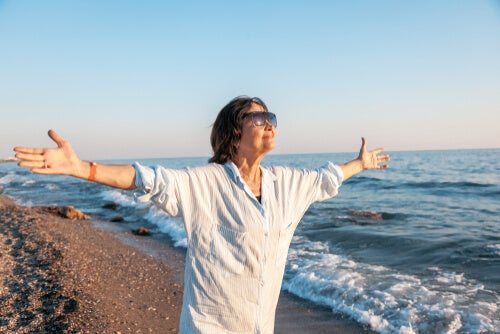 An older woman standing on a beach with her arms spread wide. 