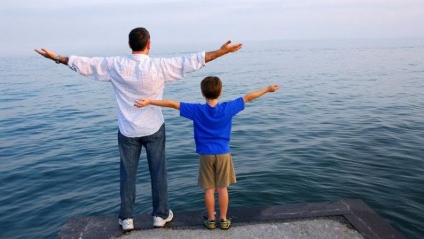Father and son with arms outstretched in front of water.
