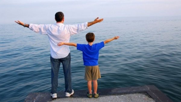 Father and son with arms outstretched in front of water.