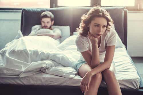 En kvinde, der sidder på kanten af seng, med mand i baggrunden