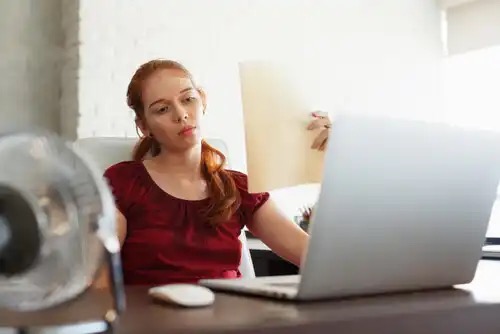 A woman working on her computer, fanning herself.
