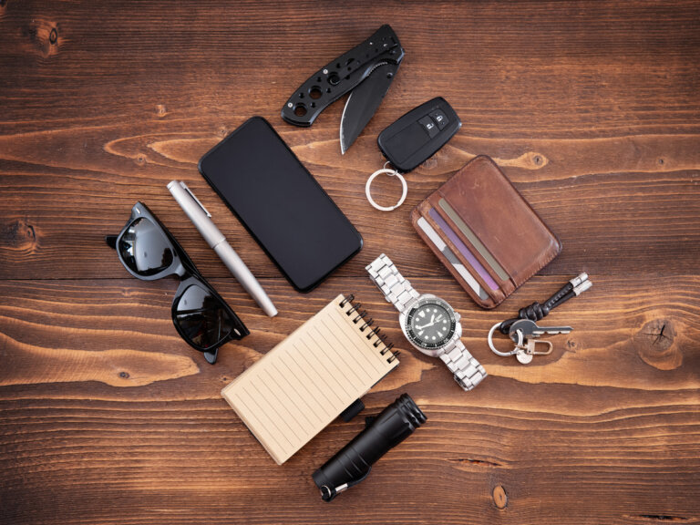 Personal items, including a wallet, cell phone, pen, watch, keys, notepad, mini flashlight, and pocket knife neatly arranged to make a square on a wooden surface.