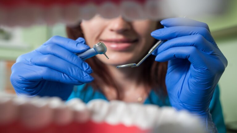 The view of a female dentist wearing blue rubber gloves and holding dental instruments from the perspective of inside the patient's mouth.