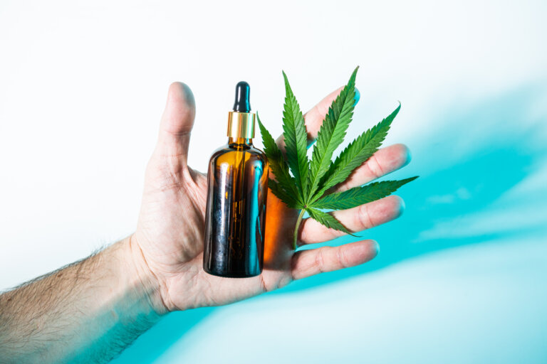 A man's hand holding a bottle of cannabis and leaves from a marijuana plant.